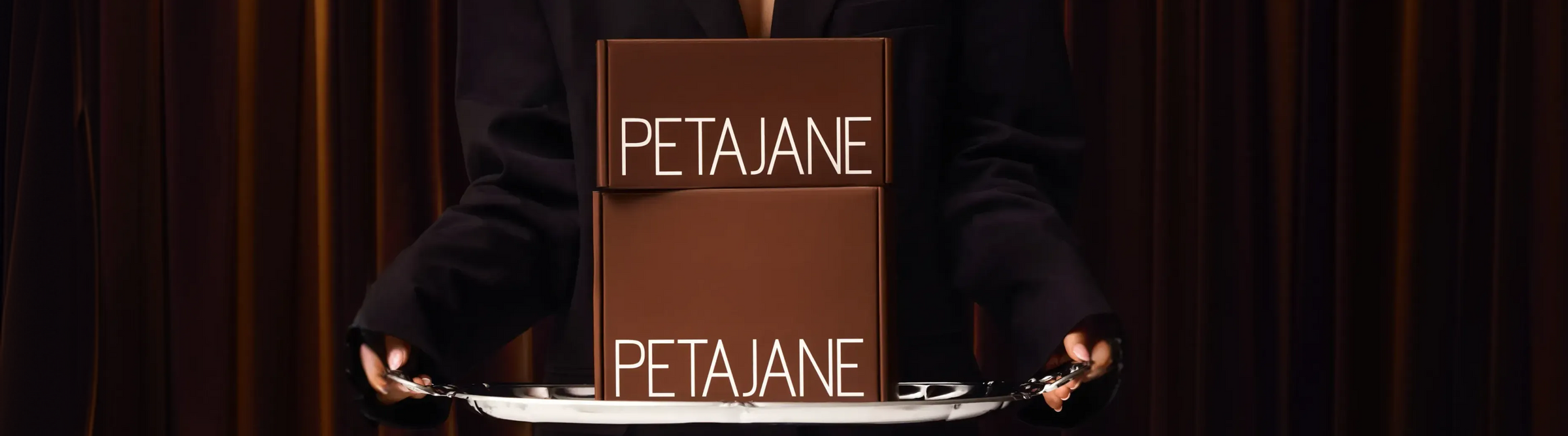 A wide shot of a woman in a black blazer holding a silver platter with stacked Peta Jane branded shipping boxes against dark curtains, highlighting a premium delivery experience.